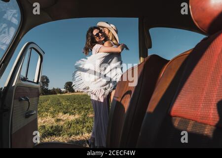 Blick von innen von Retro-Auto der Frauen umarmen und Küssen glücklich draußen gegen Landschaft mit grünen Bäumen und Himmel Stockfoto