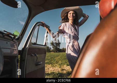 Blick von innen auf Retro-Auto der glücklichen trendigen Frau in Hut und Sonnenbrille stehen aufgeregt in der Natur und lachen Stockfoto