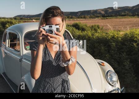 Frau in Kleid Foto mit Retro-Kamera stehen auf Hintergrund der grünen Sommerlandschaft im Sonnenlicht Stockfoto