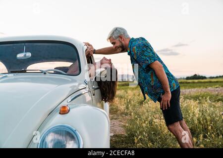 Seitenansicht einer Frau, die im Auto sitzt und sich mit dem Kopf nach außen lehnt und den Mann anschaut, der im Sommer glücklich darüber steht und lächelt Stockfoto