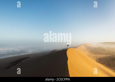 Frau, die auf einer riesigen Sanddüne in der Sahara von Marokko läuft. Schönes warmes Sonnenlicht und Nebel am Morgen. Blick von hinten. Freiheitskonzept. Stockfoto