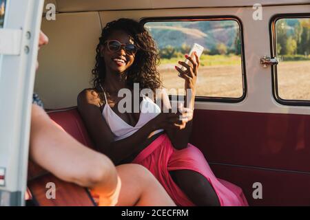 Diverse Frauen spielen akustische Gitarre und lachen, während sie zusammen sitzen Im Retro-Van während der Reise Stockfoto