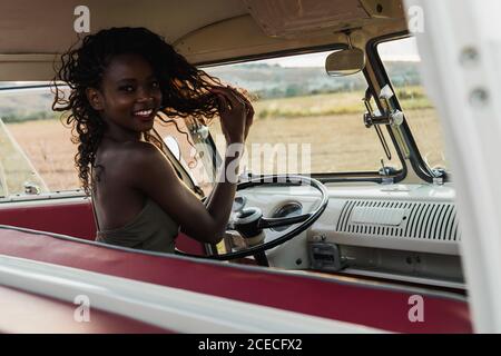Seitenansicht einer charmanten afroamerikanischen Frau, die lächelt und die Kamera anschaut, während sie auf dem Fahrersitz eines Retro-Autos sitzt und während der Fahrt auf dem Land lockiges Haar berührt Stockfoto
