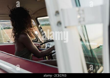 Seitenansicht einer charmanten afroamerikanischen Frau, die lächelt und die Kamera anschaut, während sie auf dem Fahrersitz eines Retro-Autos sitzt und während der Fahrt auf dem Land lockiges Haar berührt Stockfoto