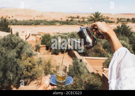 Frau Hände Gießen Glas der traditionellen Minze marokkanischen Tee mit Vintage Silber Geschirr. Sandwüste und Palmen im Hintergrund. Stockfoto