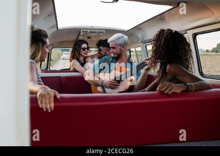 Gruppe von jungen Leuten, die lachen und auf einen gutaussehenden Kerl hören Spielen akustische Gitarre im Vintage-Van in der Natur Stockfoto
