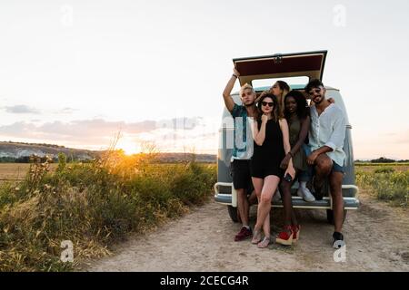 Gruppe von fröhlichen jungen Menschen in der Nähe Retro-Van und stehen Lachen, während Sie gemeinsam Zeit in der Natur verbringen Stockfoto