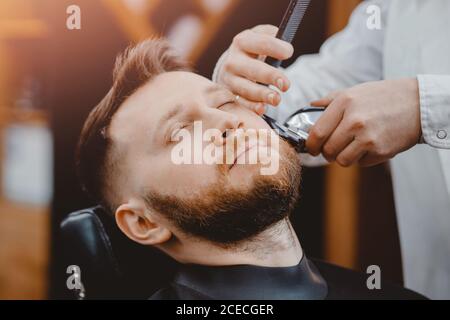 Nahaufnahme des barbers, der den Bart dem Mann in der Barbershop mit elektrischem Rasiermesser schart Stockfoto