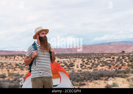 Hübscher bärtiger Mann mit Rucksack, der beim Gehen in der Nähe wegschaut Zelt an bewölktem Tag in der Wüste Stockfoto