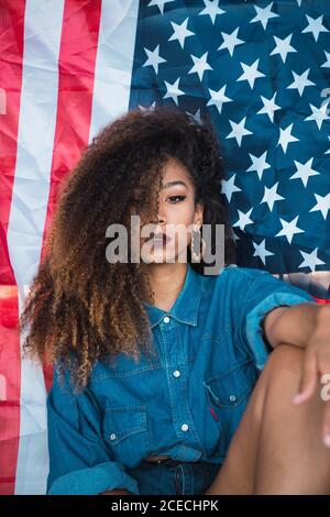 Junge afroamerikanische Frau mit lockigen Haaren und hellem Make-up im Denim-Hemd, die neben der Flagge der USA sitzt und die Kamera anschaut Stockfoto