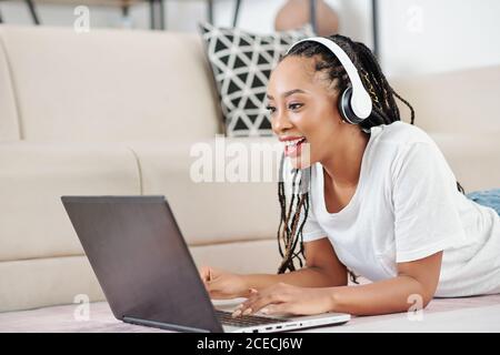 Aufgeregt schöne junge schwarze Frau in Dreadlocks liegen auf der Boden und studieren oder arbeiten auf Laptop Stockfoto