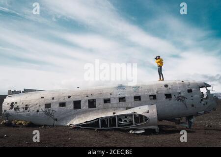 Seitenansicht des Mannes mit Fotokamera, der auf einem abgestürzten Flugzeug auf dem Boden des Geländes in Island steht. Stockfoto