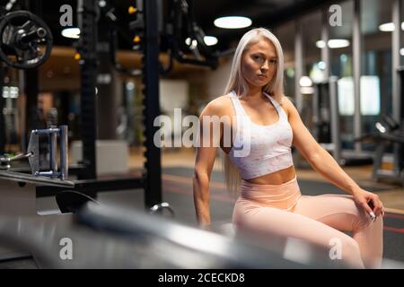 Junge schöne blonde Frau in Sportkleidung sitzt im Fitnessstudio und Blick auf die Kamera auf dem Hintergrund der Ausrüstung Stockfoto