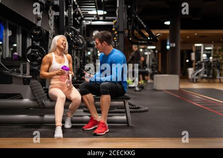 Junger hübscher Mann und blonde schöne Frau in Sportkleidung sitzen im Fitnessstudio auf Geräten und reden Stockfoto