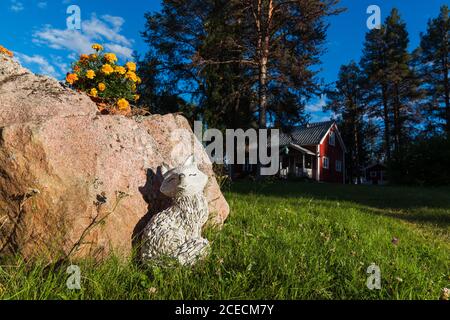 Statue des Hundes aus weißem Stein in der Nähe groß platziert Rock mit Blumen auf dem Hintergrund der Siedlung mit Holzhäuser zwischen Bäumen Stockfoto