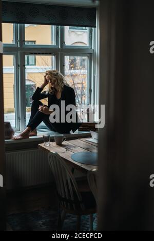 Junge, nachdenkliche Frau, die zu Hause auf dem Schweller sitzt und traurig aussieht. Stockfoto