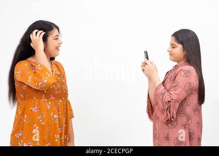 Zwei junge indische Mädchen fotografieren auf ihrem Smartphone. Nette Jugendliche, die Spaß miteinander auf einem weißen Hintergrund haben. Stockfoto