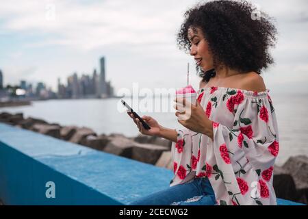 Schöne junge ethnische Frau, die Telefon benutzt, während sie ein süßes Getränk in einer Tasse am Zaun der Küste von Panama City hat Stockfoto