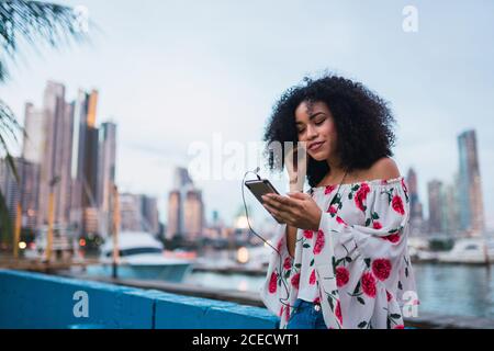 Fröhliche, moderne, ethnische Frau, die mit Kopfhörern und Smartphone am Wasser gegen die Stadtlandschaft von Panama Musik hört Stockfoto