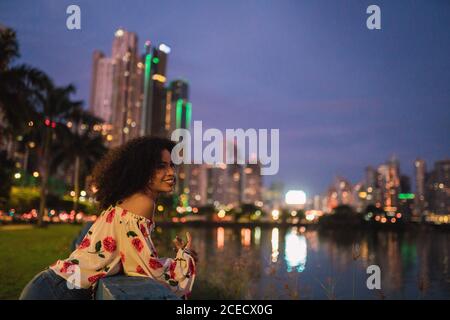 Seitenansicht einer fröhlichen schwarzen Frau am Ufer von Panama City in der Nacht mit leuchtendem Stadtbild im Hintergrund Stockfoto