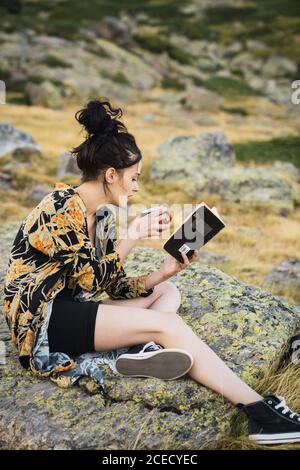 Seitenansicht einer jungen Frau, die eine Tasse Kaffee auf einem Felsen im Tal sitzt und ein Buch liest Stockfoto