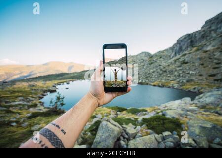 Crop tätowierte männliche Hand hält Smartphone mit Bild einer Frau auf dem Felsen stehen am Ufer des Bergsees Stockfoto