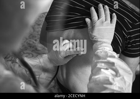 Beschnittene Nahaufnahme der Hand des Arztes in einem PSA-Schutzanzug, bei dem ein älterer männlicher Patient zu Hause mit einem Phonendoskop untersucht und dabei seinen Rücken überprüft wird. Schwarzweiß-Bild. COVID-19 Konzept Stockfoto