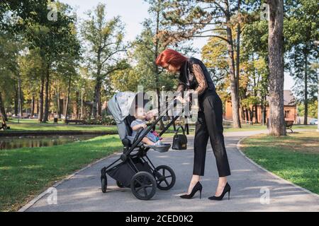 Seitenansicht der schönen Frau im eleganten Outfit stehen auf park Gasse in der Nähe Kinderwagen mit niedlichen Baby Mädchen Stockfoto