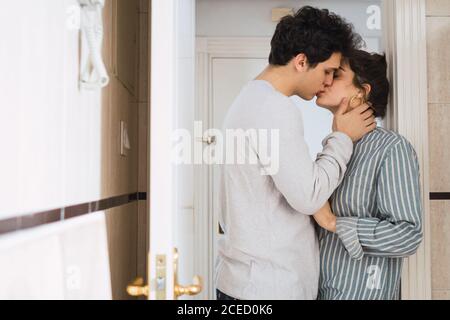 Seitenansicht des jungen Paares im Pyjama stehend und küssend in der Tür zu Hause. Stockfoto