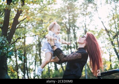 Seitenansicht der hübschen Dame mit Ingwer Haar hält Baby Mädchen und spielen mit ihr, während sie auf verschwommenem Hintergrund stehen Von Parkbäumen am sonnigen Tag Stockfoto