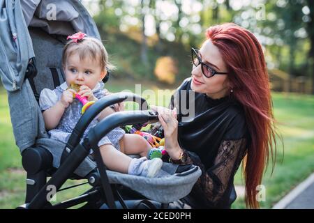 Attraktive Frau mit Ingwer-Haar lächelnd und Blick auf entzückende Baby-Mädchen mit Spielzeug in der Kutsche auf verschwommenem Hintergrund†of Park Stockfoto