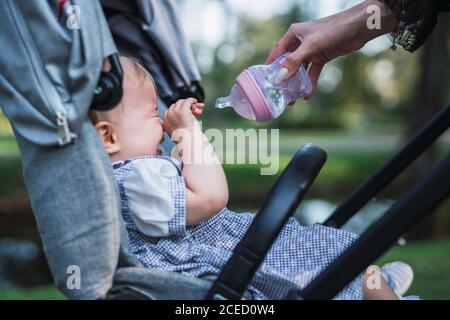 Hand der anonymen Mutter hält Flasche Wasser und versuchen Zu trösten weinendes Baby im Kinderwagen auf verschwommenem Hintergrund parken Stockfoto