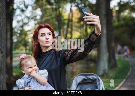 Schöne junge Frau mit Ingwerhaar hält niedliches Baby Mädchen und posiert für Selfie, während sie auf verschwommenem Hintergrund des Parks steht Stockfoto