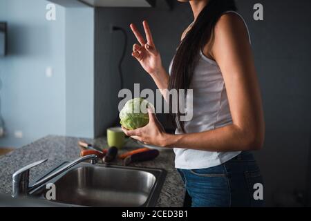 Schöne leidenschaftliche Brünette Hispanic Frau Waschen Kohl in Waschbecken in der Nähe von Karotten, Auberginen und Tasse in der modernen Küche Stockfoto