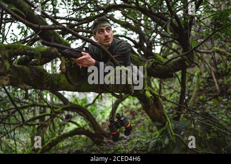 Soldat oder revolutionäres Mitglied oder Jäger in Tarnung auf dem Baum, der die Waffe in der Hand beobachtet Stockfoto