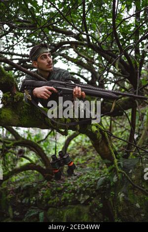 Soldat oder revolutionäres Mitglied oder Jäger in Tarnung auf dem Baum, der die Waffe in der Hand beobachtet Stockfoto