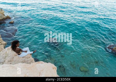 Von oben Aufnahme einer schwarzen Frau, die sich allein auf einer Klippe über dem türkisfarbenen Wasser des Meeres entspannt Stockfoto
