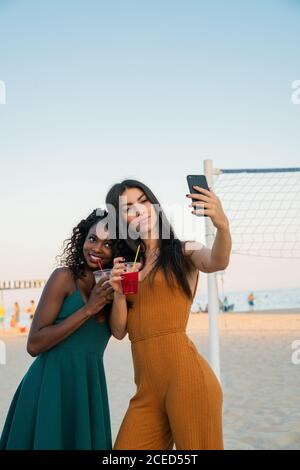 Stylische, vielfältige Frauen mit Getränken, die Selfie am Strand machen Stockfoto