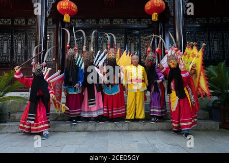 GUINZHOU, CHINA - 14. JUNI 2018: Gruppe der ethnischen Minderheit Miao Frauen in hellen traditionellen Kostümen und Masken stehen mit Mann in gelben Kostümen im Freien an der Wand des chinesischen Gebäudes in Guizhou Stockfoto