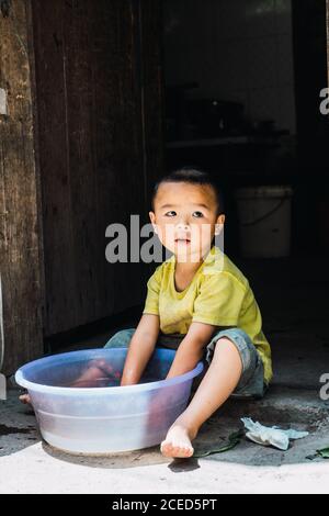 GUINZHOU, CHINA - 14. JUNI 2018: Entzückendes kleines Kind der ethnischen Minderheit Miao in Sommerkleidung sitzt auf Steinboden mit Händen in Plastikbecken mit Wasser und schaut weg in der Provinz Guizhou in China Stockfoto