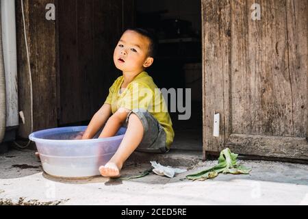 GUINZHOU, CHINA - 14. JUNI 2018: Entzückendes kleines Kind der ethnischen Minderheit Miao in Sommerkleidung sitzt auf Steinboden mit Händen in Plastikbecken mit Wasser und schaut weg in der Provinz Guizhou in China Stockfoto