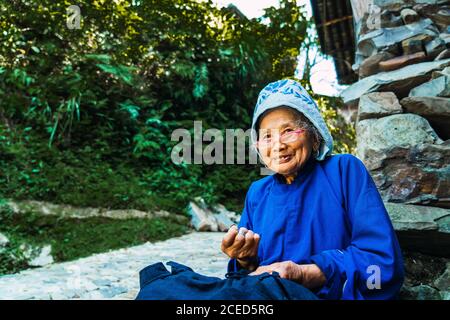 GUINZHOU, CHINA - 14. JUNI 2018: Seitenansicht einer alten chinesischen Frau in einer Brille und traditionellen blauen Kleidern und einer Mütze, die in den Händen genäht wurde und mit einer anderen Frau aus der Miao-Ethnie und Bäumen auf unscharfem Hintergrund auf Steinen sitzend in Guizhou, China Stockfoto