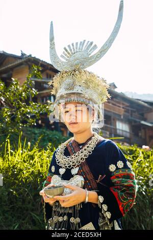 GUINZHOU, CHINA - 14. JUNI 2018: Attraktive junge chinesische Frau der ethnischen Minderheit Miao, die in traditionellen Kleidern mit schönen Ornamenten und silbernem Horn-Kopfschmuck auf dem Bürgersteig des Parks läuft und in der Provinz Guizhou lächelt Stockfoto