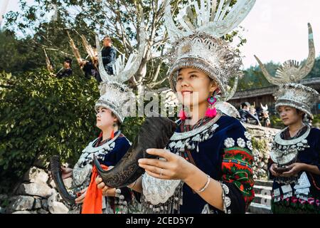 GUINZHOU, CHINA - 14. JUNI 2018: Attraktive junge chinesische Frauen der ethnischen Minderheit Miao, die in traditionellen Kleidern mit schönen Ornamenten und silbernen Horn Kopfschmuck auf dem Parksteig spazieren und in der Provinz Guizhou lächeln Stockfoto