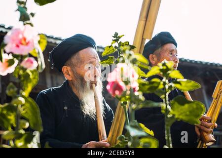 GUINZHOU, CHINA - 14. JUNI 2018: Ältere Männer in dunklen ethnischen Kostümen von Miao Menschen, die traditionelle Bambusflöten spielen, stehen am Sommertag im Freien beim Bauen mit hellen Blumen im Vordergrund in der chinesischen Provinz Guizhou Stockfoto