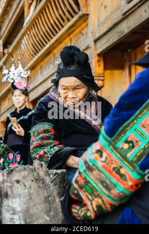 GUINZHOU, CHINA - 14. JUNI 2018: Lächelnde ältere Frau in dunklem Kostüm mit lokalen Ornamenten, die mit einer Gruppe anderer Frauen ihrer ethnischen Gruppe in einem alten Holzgebäude steht und lächelt in der chinesischen Provinz Guizhou Stockfoto