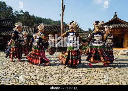 GUINZHOU, CHINA - 14. JUNI 2018: Frauen der Miao Ethnie in hellen Kostümen mit bunten Ornamenten und Kopfschmuck Durchführung traditionellen Festtanz auf Dorfplatz an sonnigen Sommertag in der Provinz Guizhou in China Stockfoto