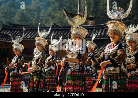 GUINZHOU, CHINA - 14. JUNI 2018: Frauen der Miao Ethnie in hellen Kostümen mit bunten Ornamenten und Kopfschmuck Durchführung traditionellen Festtanz auf Dorfplatz an sonnigen Sommertag in der Provinz Guizhou in China Stockfoto