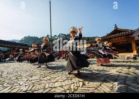 GUINZHOU, CHINA - 14. JUNI 2018: Frauen der Miao Ethnie in hellen Kostümen mit bunten Ornamenten und Kopfschmuck Durchführung traditionellen Festtanz auf Dorfplatz an sonnigen Sommertag in der Provinz Guizhou in China Stockfoto