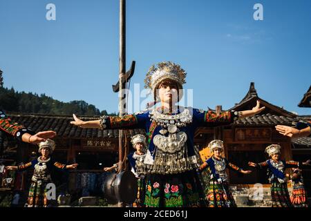 GUINZHOU, CHINA - 14. JUNI 2018: Frauen der Miao Ethnie in hellen Kostümen mit bunten Ornamenten und Kopfschmuck Durchführung traditionellen Festtanz auf Dorfplatz an sonnigen Sommertag in der Provinz Guizhou in China Stockfoto